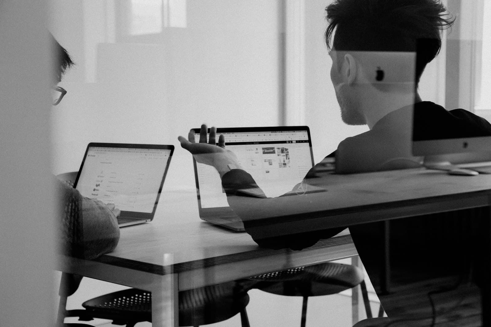 Two people reviewing work on laptops, photographed in black and white.